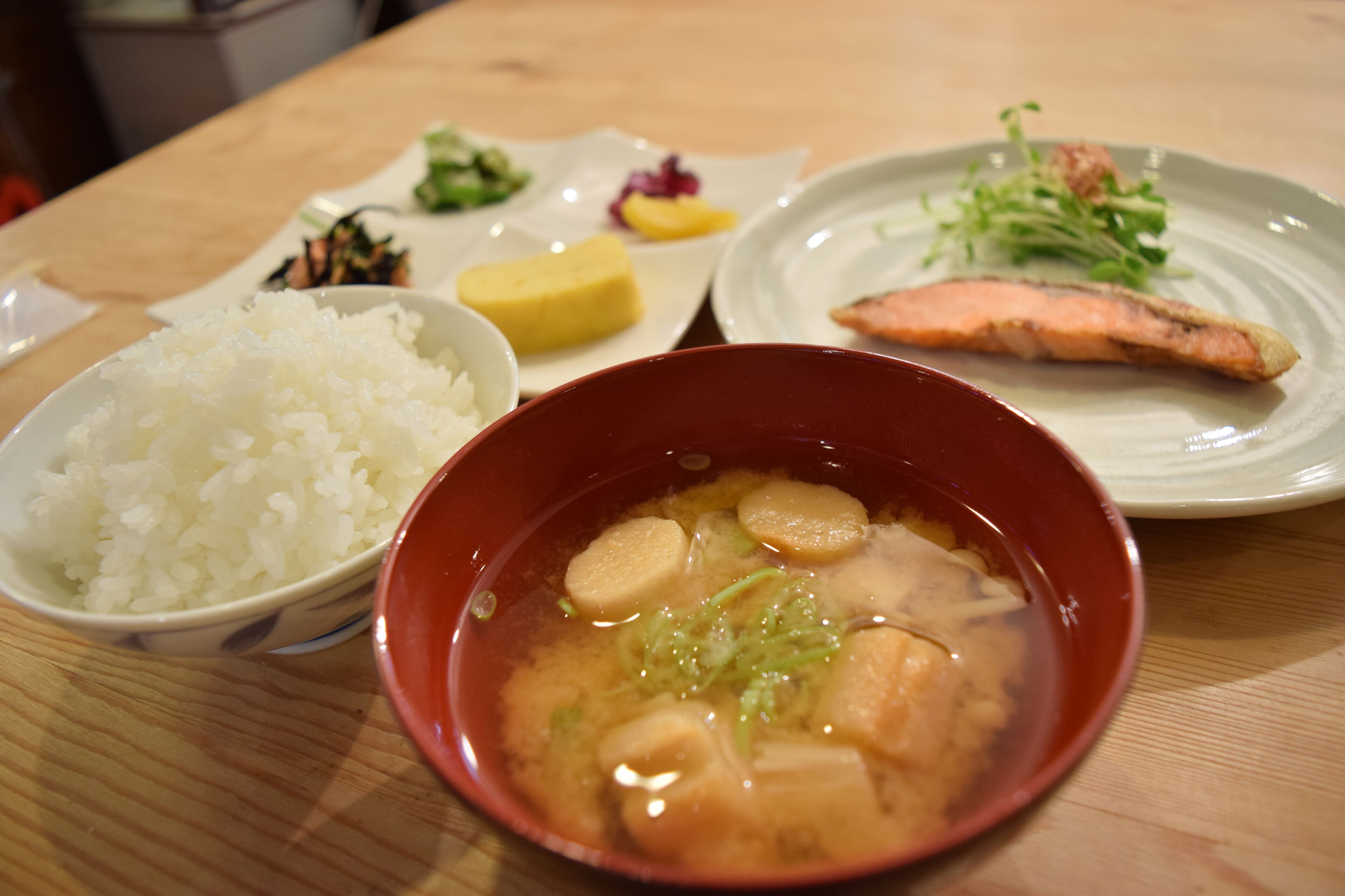 Una mesa con un tazón de arroz, un tazón de sopa, un plato con un trozo de salmón y un plato con una pequeña guarnición de vegetales encurtidos.