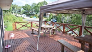 A wooden deck with picnic tables and benches, a covered area, and a view of the street and greenery.