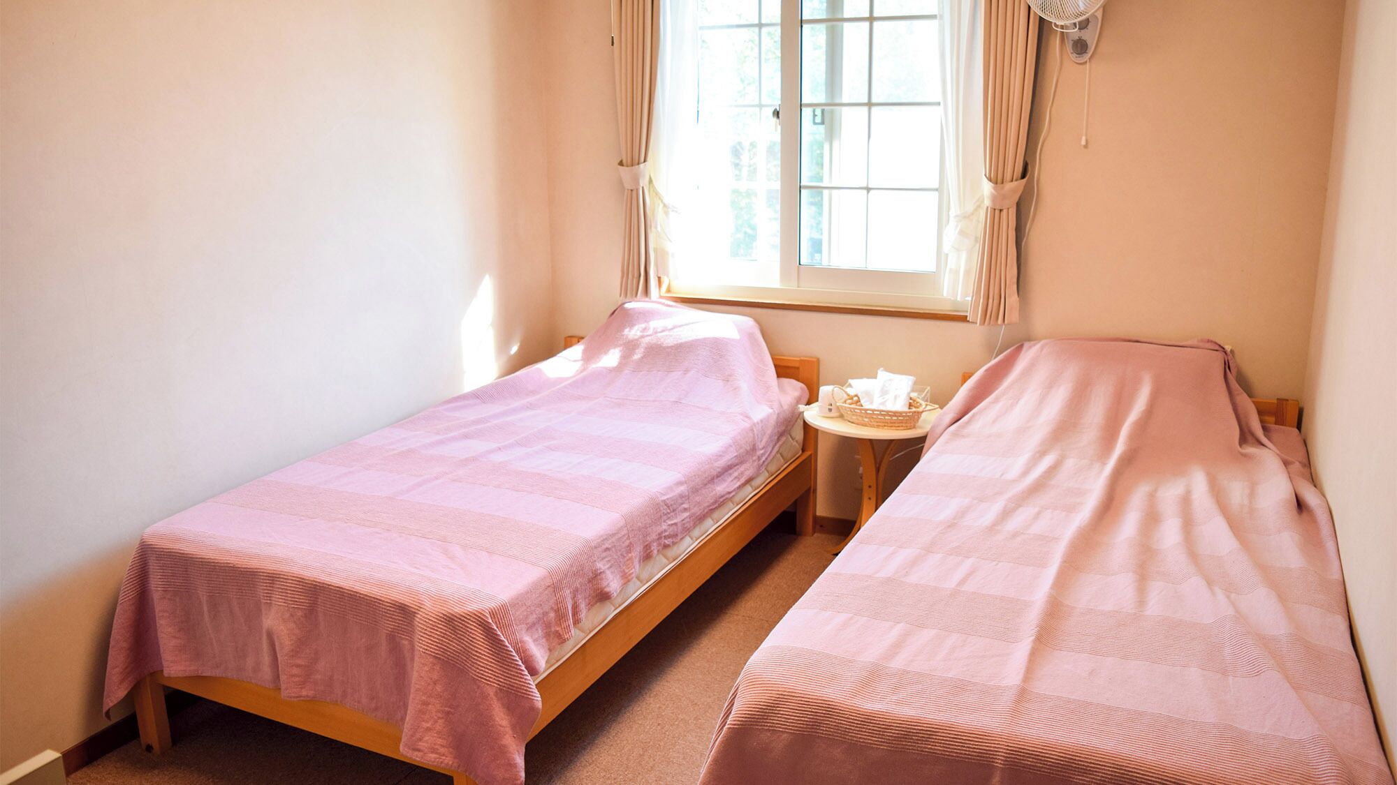 Una habitación pequeña y luminosa con dos camas individuales, una mesita de noche con una cesta, y una ventana con cortinas.
