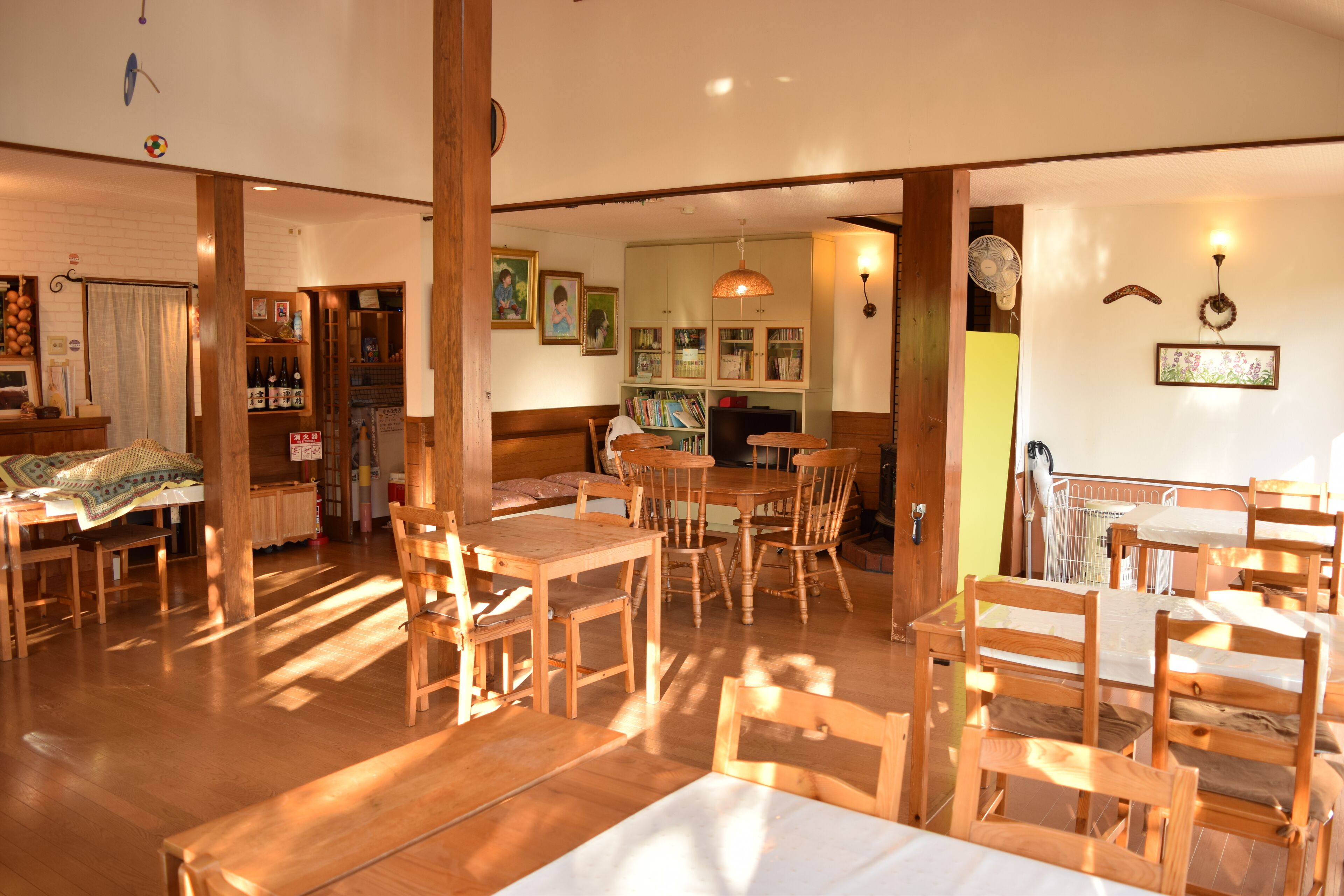 A spacious dining area with wooden tables and chairs, a kitchen in the background, and a bookshelf filled with books.
