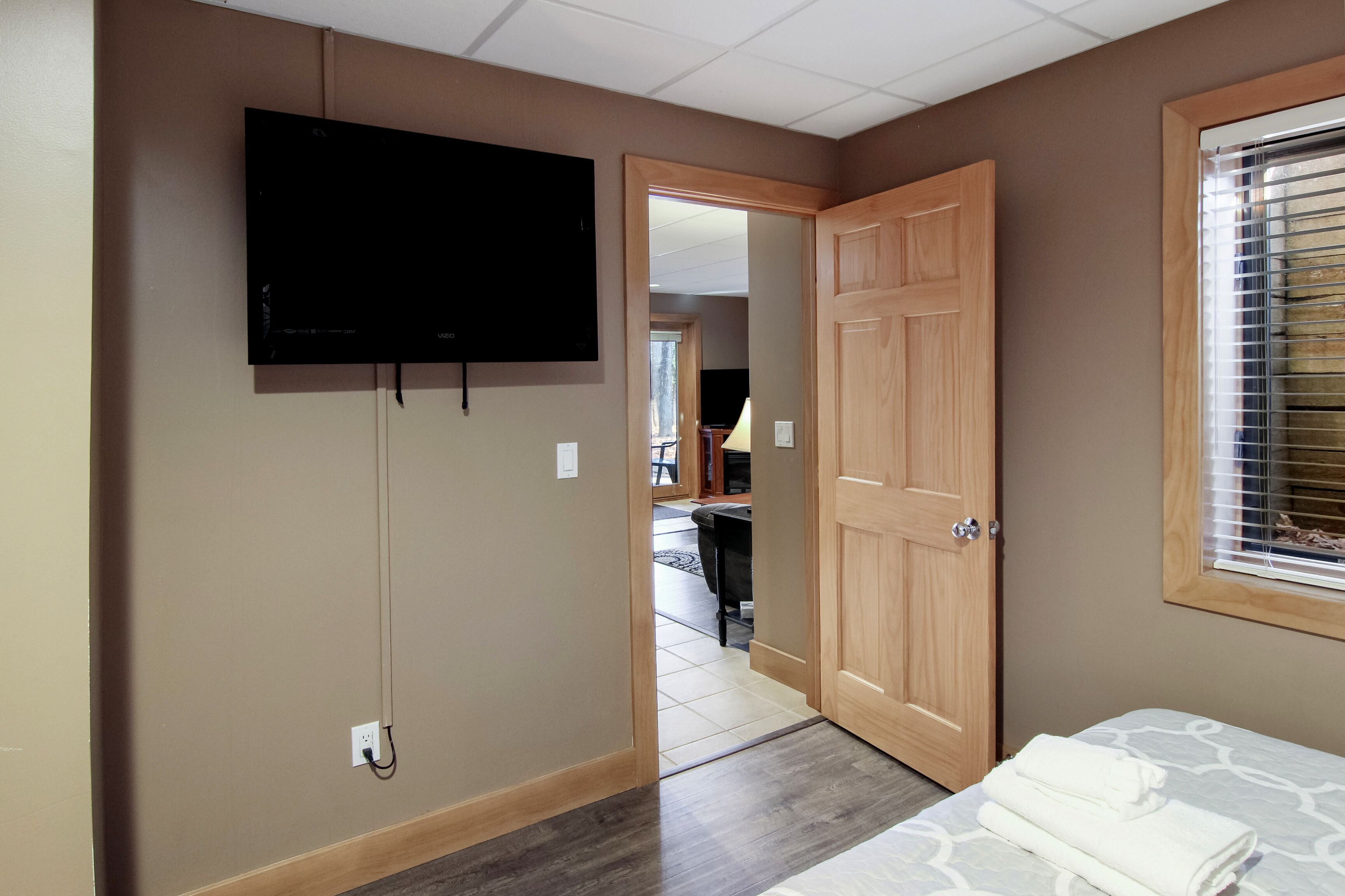 A hotel room with a television mounted on the wall, a wooden door, a window with blinds, and a bed with a patterned bedspread.