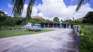 A boat docked near a covered structure with a concrete pathway leading to it, surrounded by greenery and hills in the background.