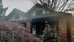 A two-story house with a brick chimney, a porch with a yellow light, and a window with a view of the surrounding area. - 3BR 2BA Dynamite Cooper Young Craftsman 2 blocks from Cooper Young intersection! (Memphis)