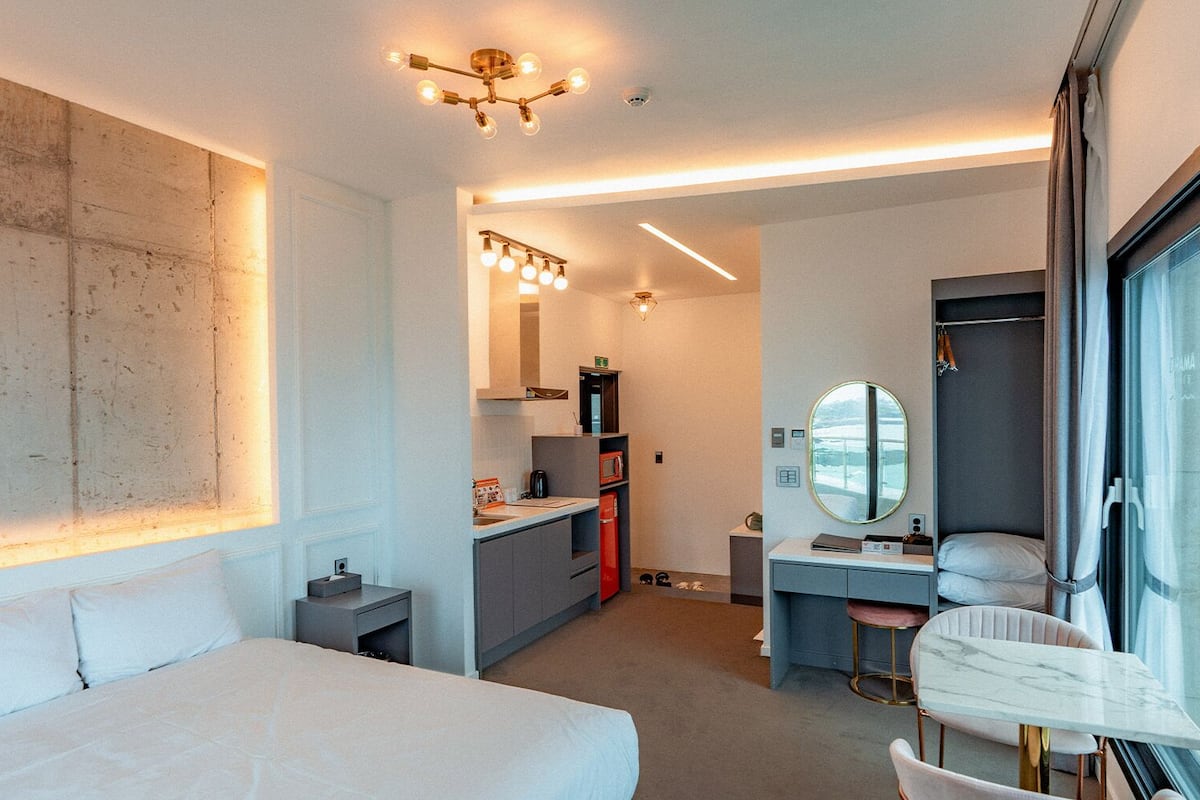 Una habitación de hotel moderna con una cama grande, un escritorio con espejo, una kitchenette y un baño con ducha.
