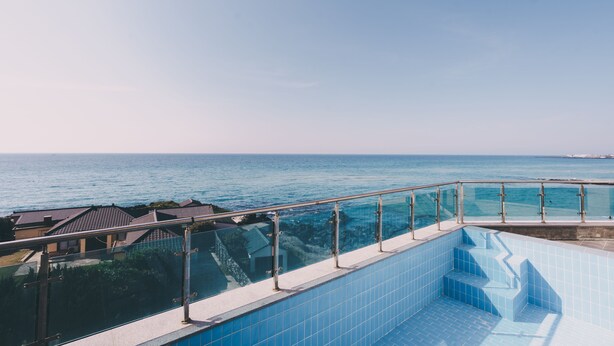 Una piscina amb una tanca de vidre que domina el mar, un cel serè i edificis residencials al fons.