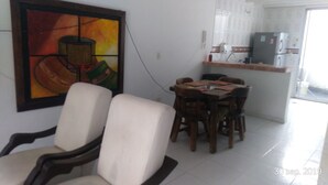 A dining area with a table and chairs, a painting on the wall, and a kitchen in the background.
