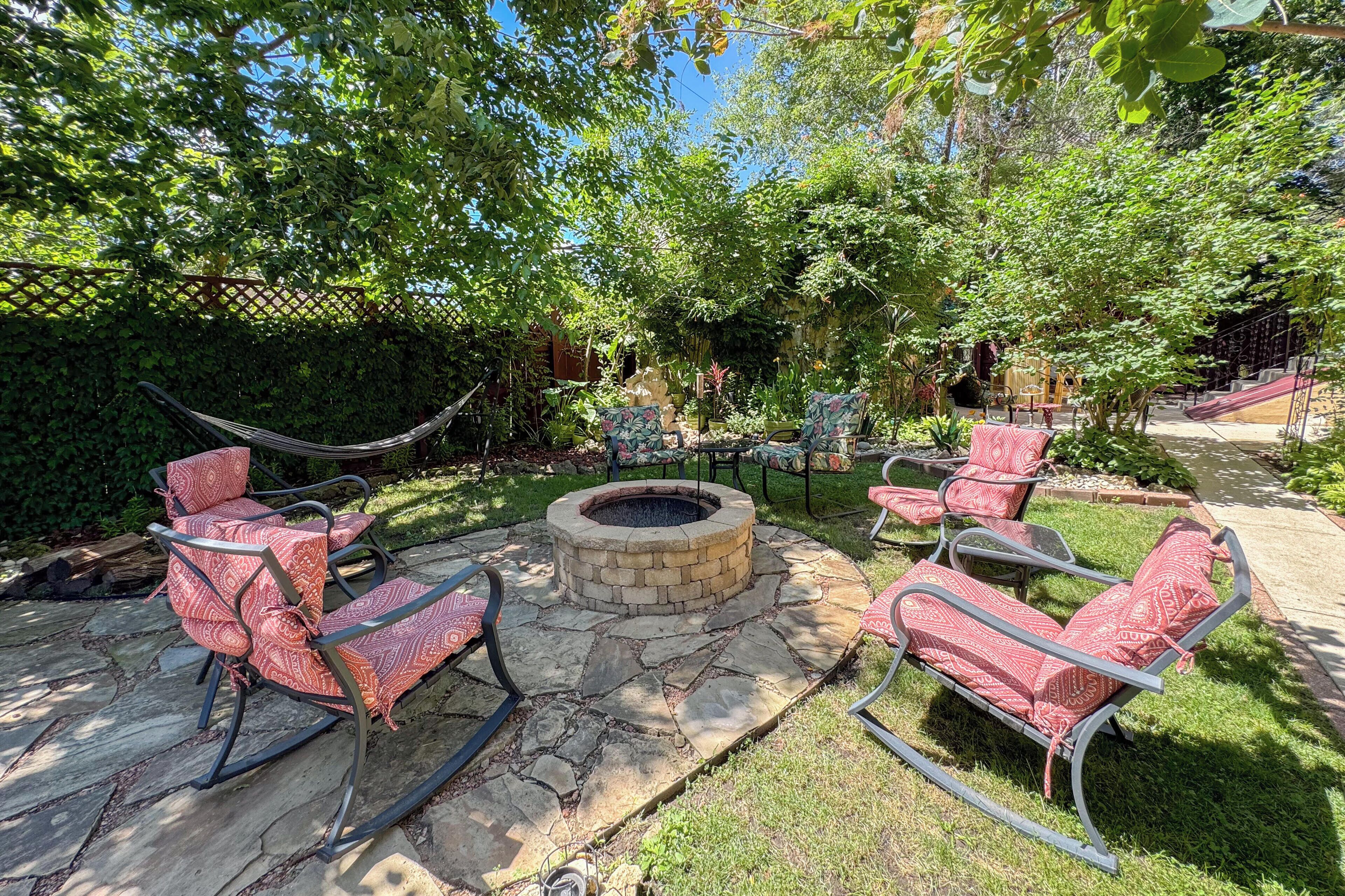 A backyard with a stone fire pit, red patterned chairs, and a hammock.