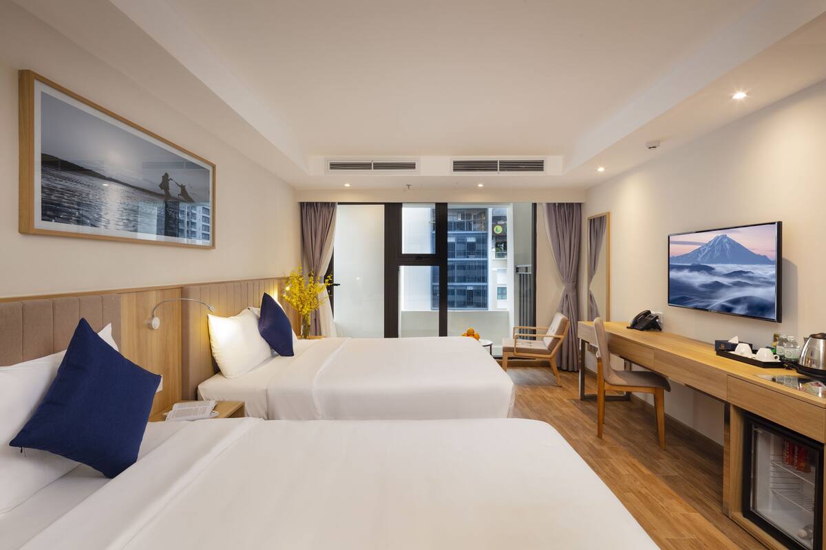 Habitació d'hotel amb dos llits, escriptori, cadira, televisió i finestra amb vista a edificis.