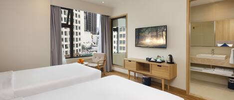 Habitación de hotel con dos camas, un escritorio, una silla, un ventanal con cortinas, un cuadro y un baño con espejo y lavamanos.