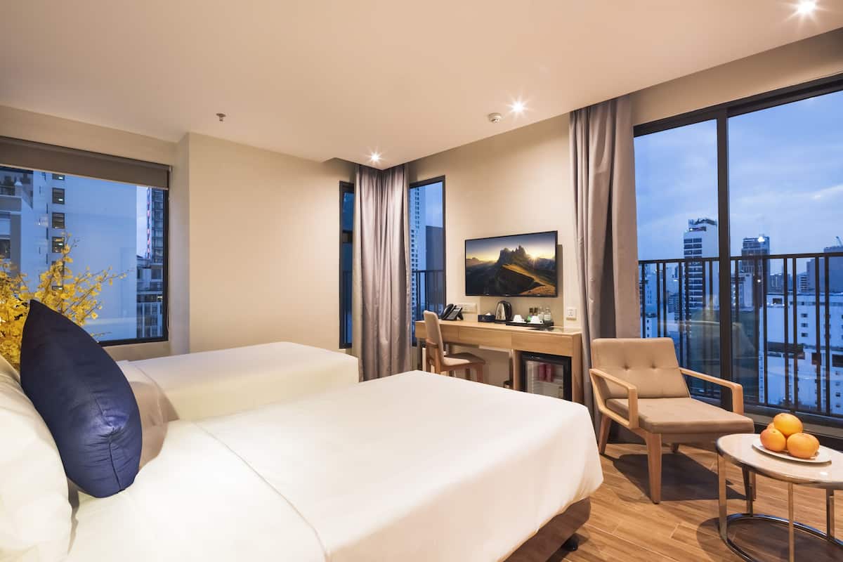 Habitació d'hotel amb dos llits, una escriptori, una cadira, un televisor i un balcó amb vista a la ciutat.