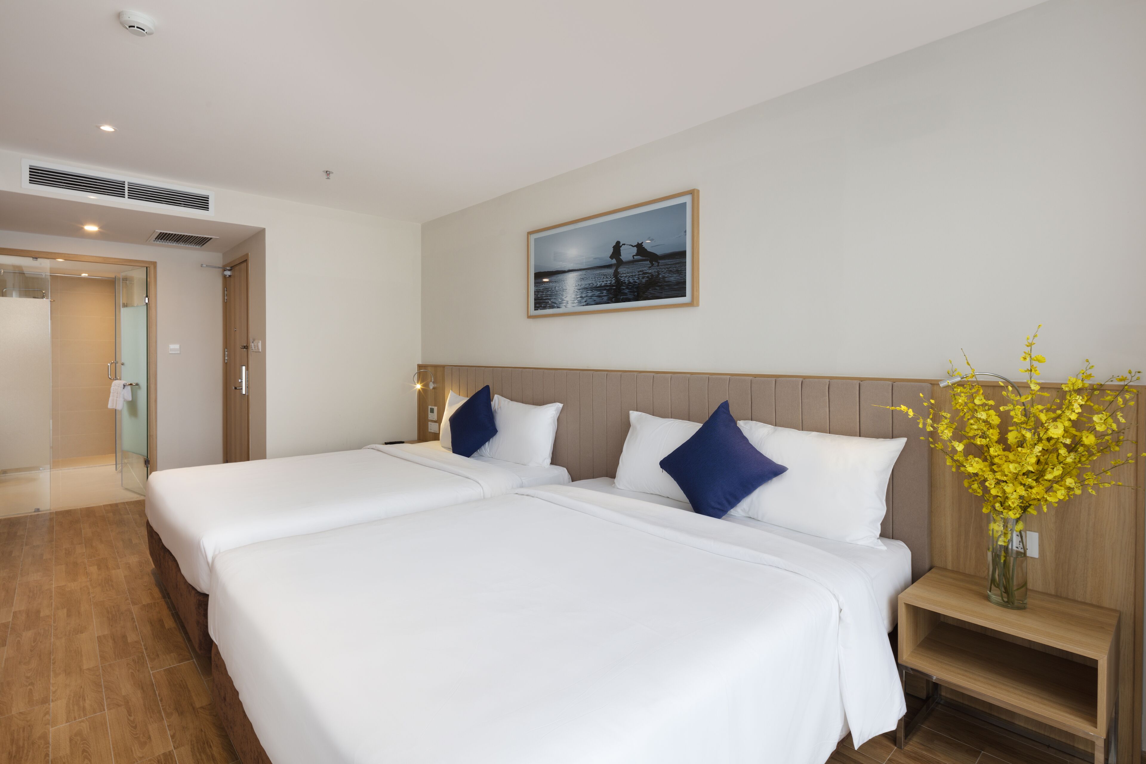 A hotel room with a large bed, a bedside table, a nightstand, a framed picture on the wall, and a vase with flowers.