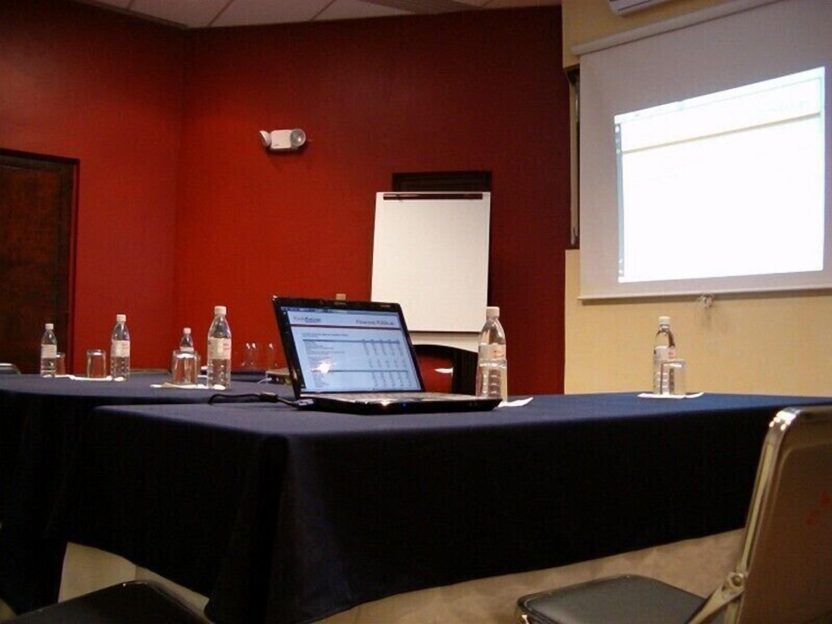 Uma sala de conferência com uma mesa comprida, laptops, garrafas de água e uma tela de projetor.