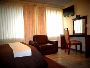 A hotel room with a bed, a brown leather chair, a wooden desk with a chair, and a television mounted on the wall.