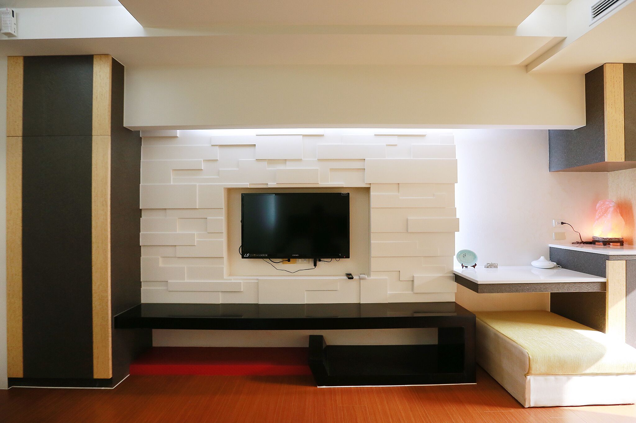 A modern living room with a flat-screen TV mounted on a white brick wall, a black entertainment unit, and a small seating area with a red accent wall.