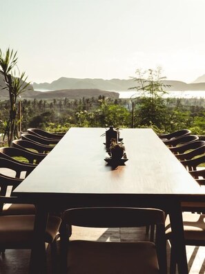 A long dining table with chairs, a bottle, and a candle, set against a backdrop of a scenic view with mountains and greenery. - Villa Strata (Kuta)