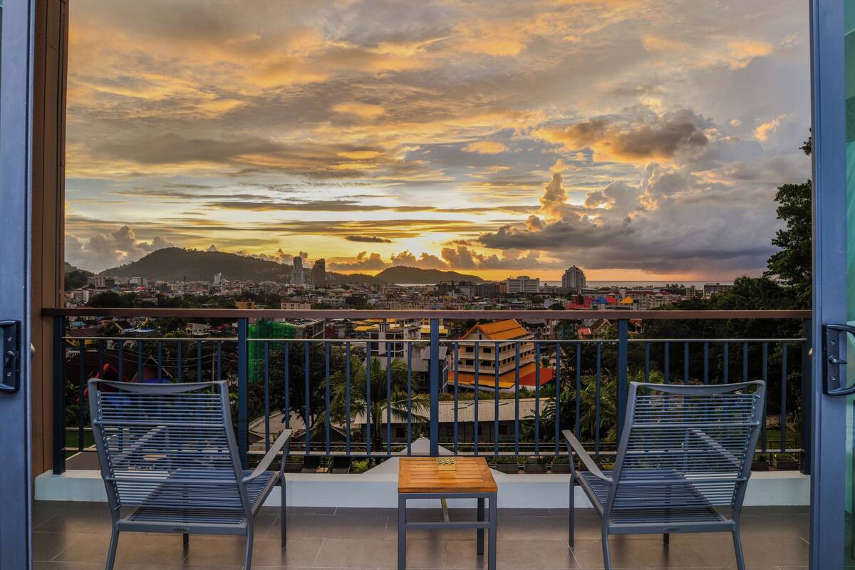 Ein Balkon mit zwei Stühlen und einem Tisch, mit Blick auf eine Stadtlandschaft mit Gebäuden und einem Hügel.