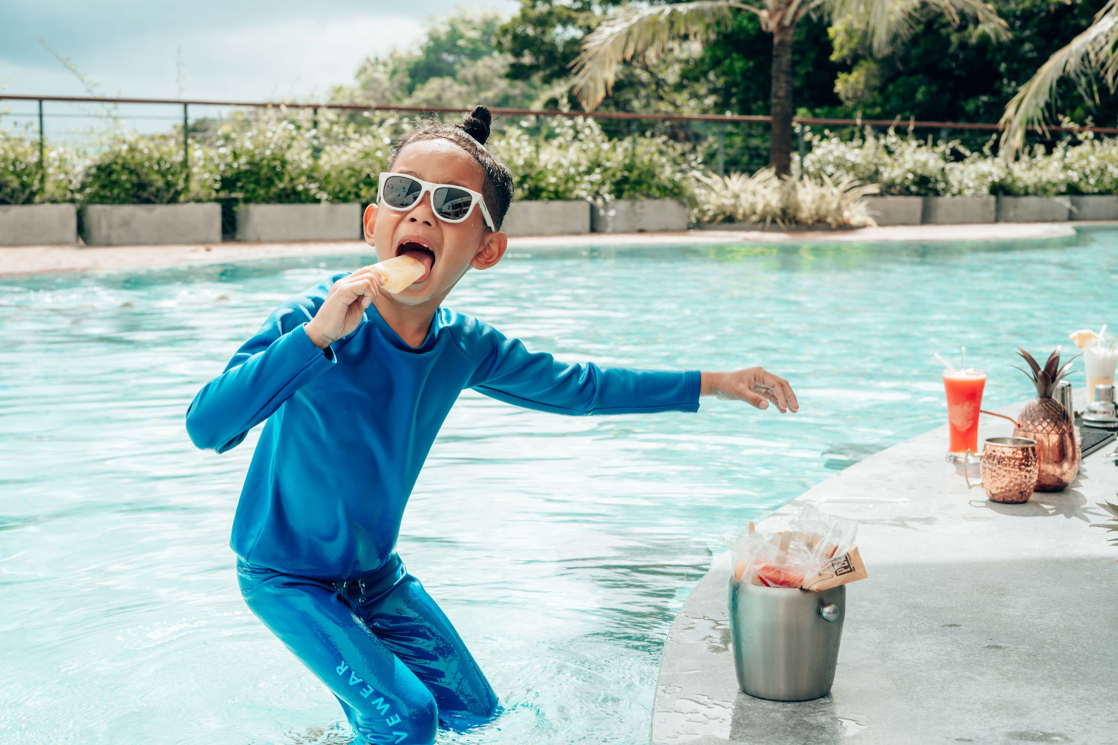 Une personne vêtue d’un maillot de bain bleu, près d’un bar au bord de la piscine, tenant un verre et un ananas.
