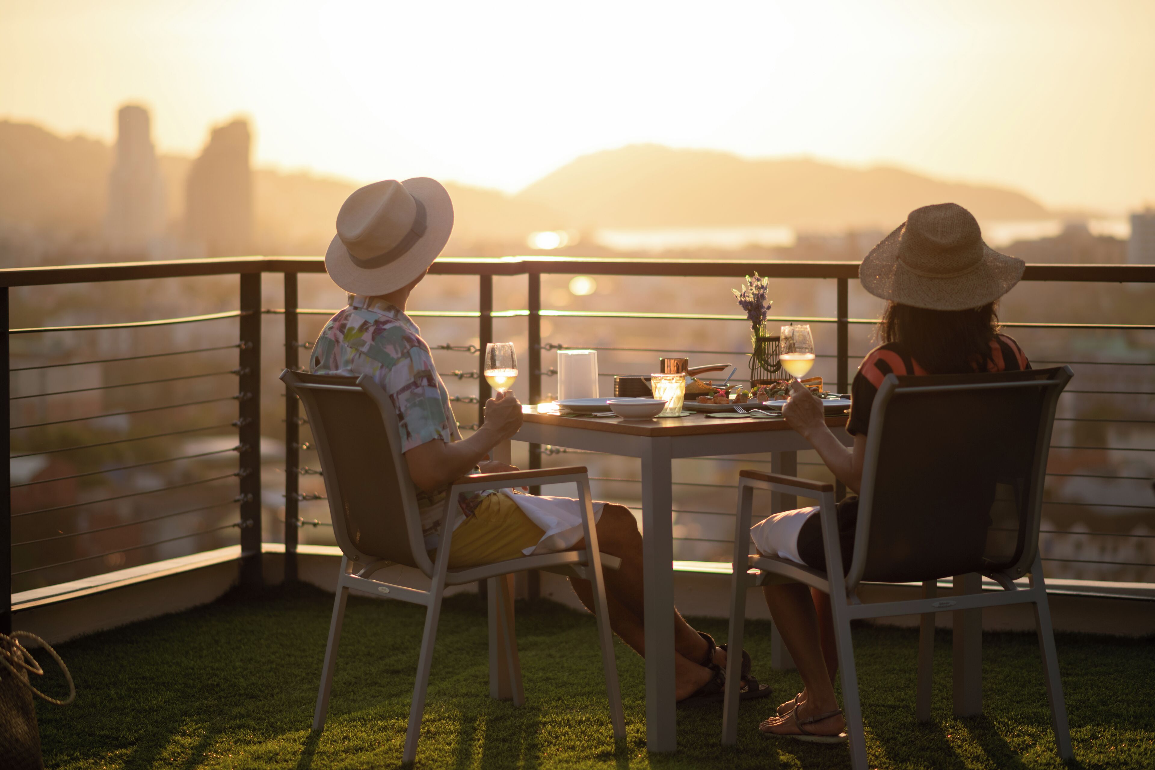 Dos personas cenando en una mesa al aire libre con vista a la ciudad, una barandilla y una zona ajardinada.