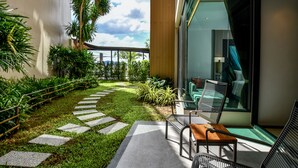 Een modern terras buiten, met een betonnen pad, zithoek en weelderige begroeiing.
