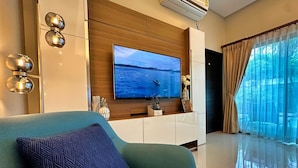 A modern living room with a large flat-screen TV mounted on a wooden wall, a sofa with a patterned cushion, a white cabinet below the TV, and a large window with sheer curtains.