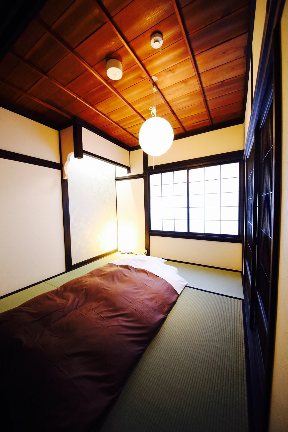 Una habitación tradicional japonesa con suelo de tatami, un futón bajo, un shoji y un techo con vigas de madera y una lámpara colgante.