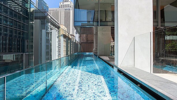 A modern indoor swimming pool with clear blue water, surrounded by high-rise buildings and glass railings.