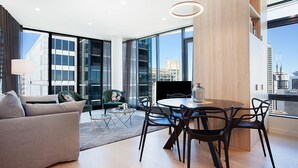 A modern living room with a sofa, chairs, a coffee table, and a dining area with a TV. Large windows offer a view of the cityscape.