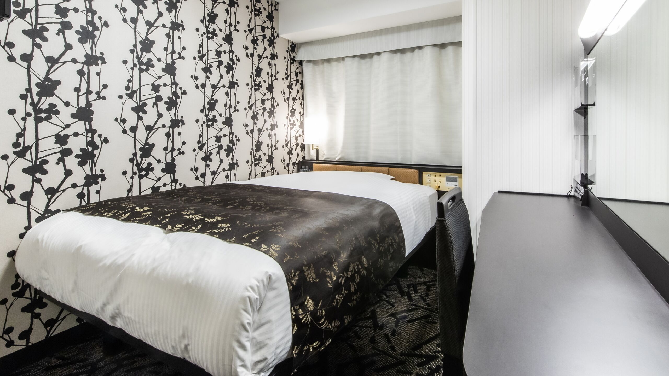 Habitación de hotel con cama, escritorio, silla, ventana con cortinas y pared con estampado floral.