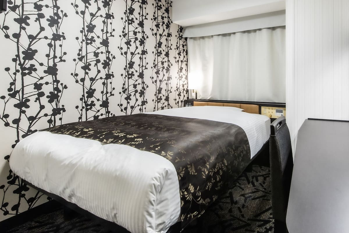 Une chambre d’hôtel comprenant un lit, un bureau, une chaise, une fenêtre avec des rideaux et un mur orné d’un motif floral.