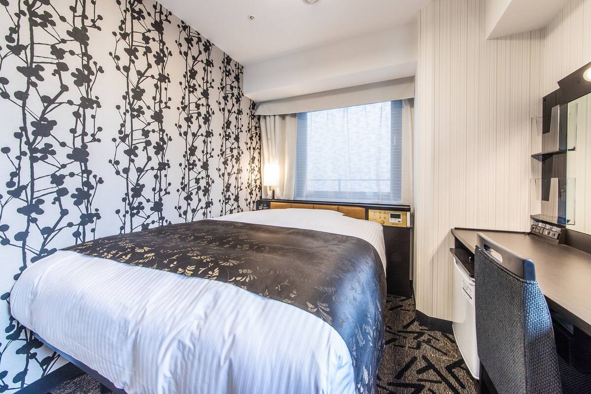 Une chambre d’hôtel comprenant un lit, un bureau, une chaise, une fenêtre avec des rideaux et un mur orné d’un motif floral.