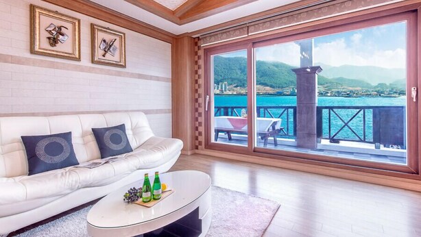 Ein modernes Wohnzimmer mit einem weißen Sofa, zwei gerahmten Kunstwerken und einem Glastisch mit Flaschen und Trauben. Eine große Schiebeglas-Tür führt auf einen Balkon mit Blick auf das Meer und die Berge.