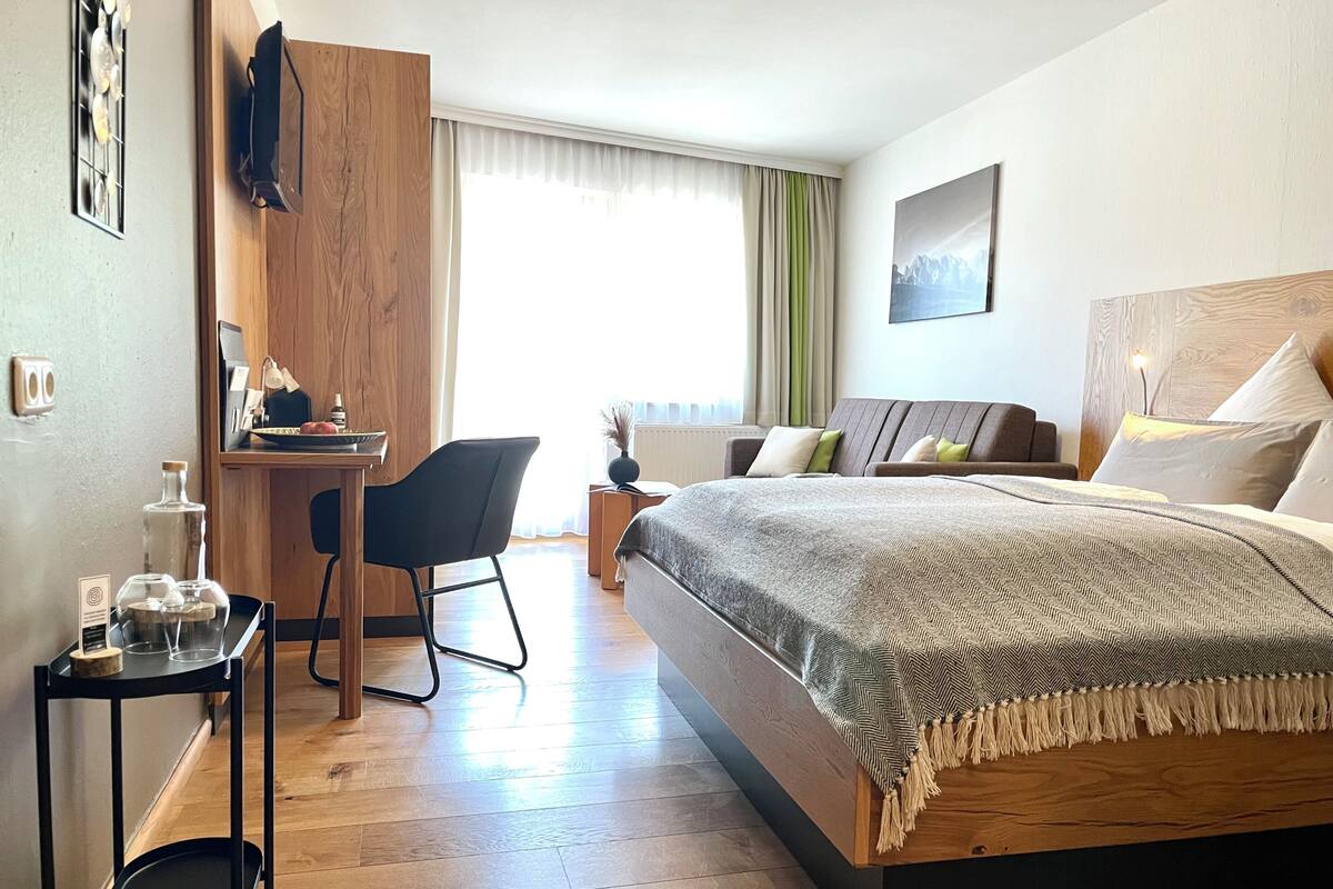Ein modernes Hotelzimmer mit einem hölzernen Bett, einem Schreibtisch mit Stuhl, einem kleinen Tisch mit Lampe und einem großen Fenster mit Vorhängen.