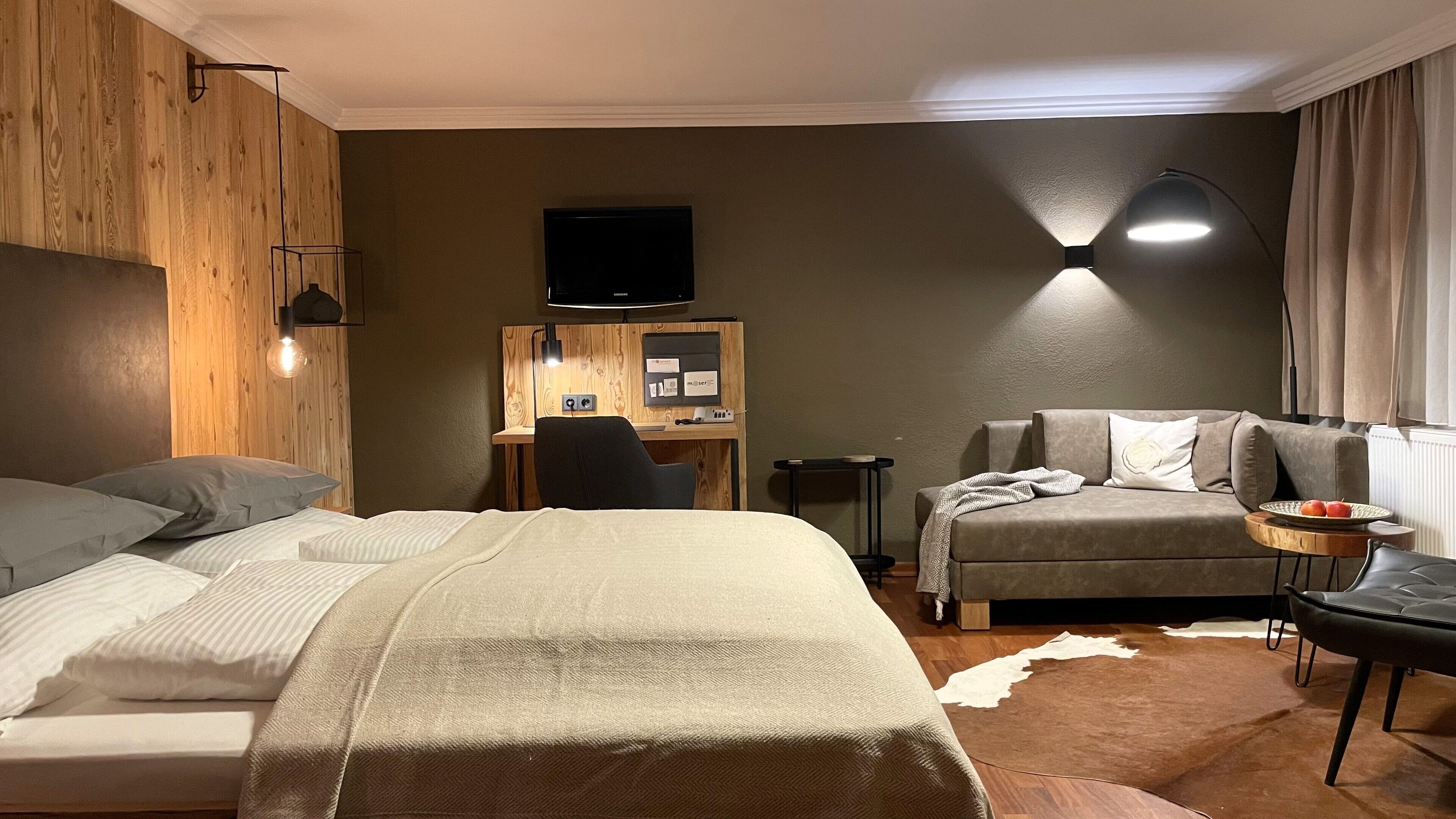 A hotel room with a bed, a sofa, a desk with a computer, a TV, and a small table with apples.