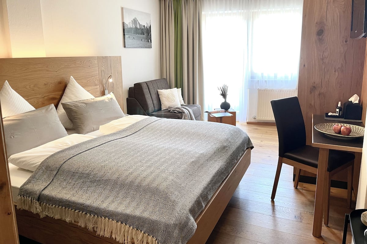Ein Hotelzimmer mit einem großen Bett, einem Schreibtisch mit Stuhl, einem kleinen Tisch mit Vase und einem Fenster mit Vorhängen.