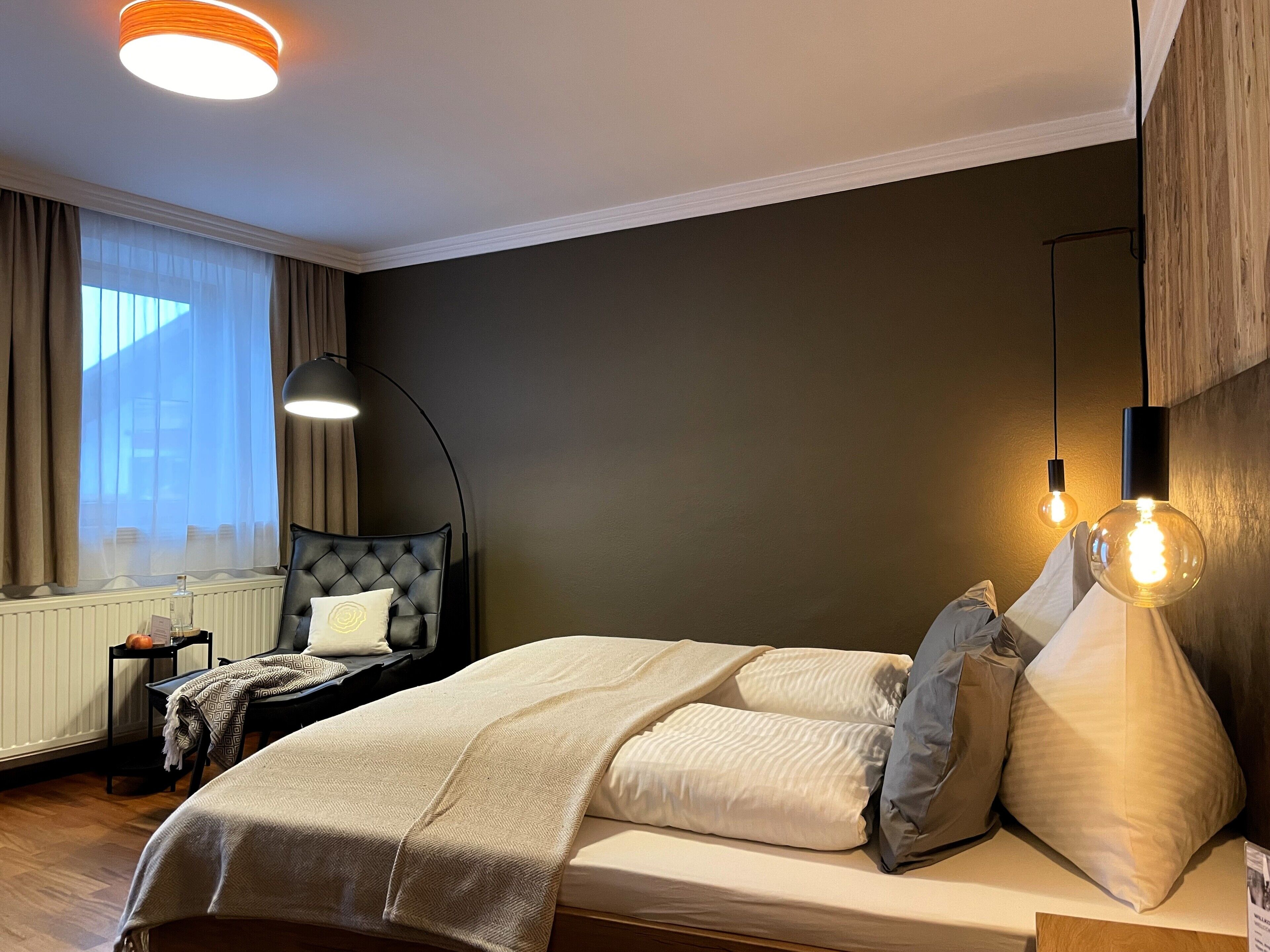 A modern bedroom with a large bed, a nightstand, a lamp, and a view of the outdoors through a window.