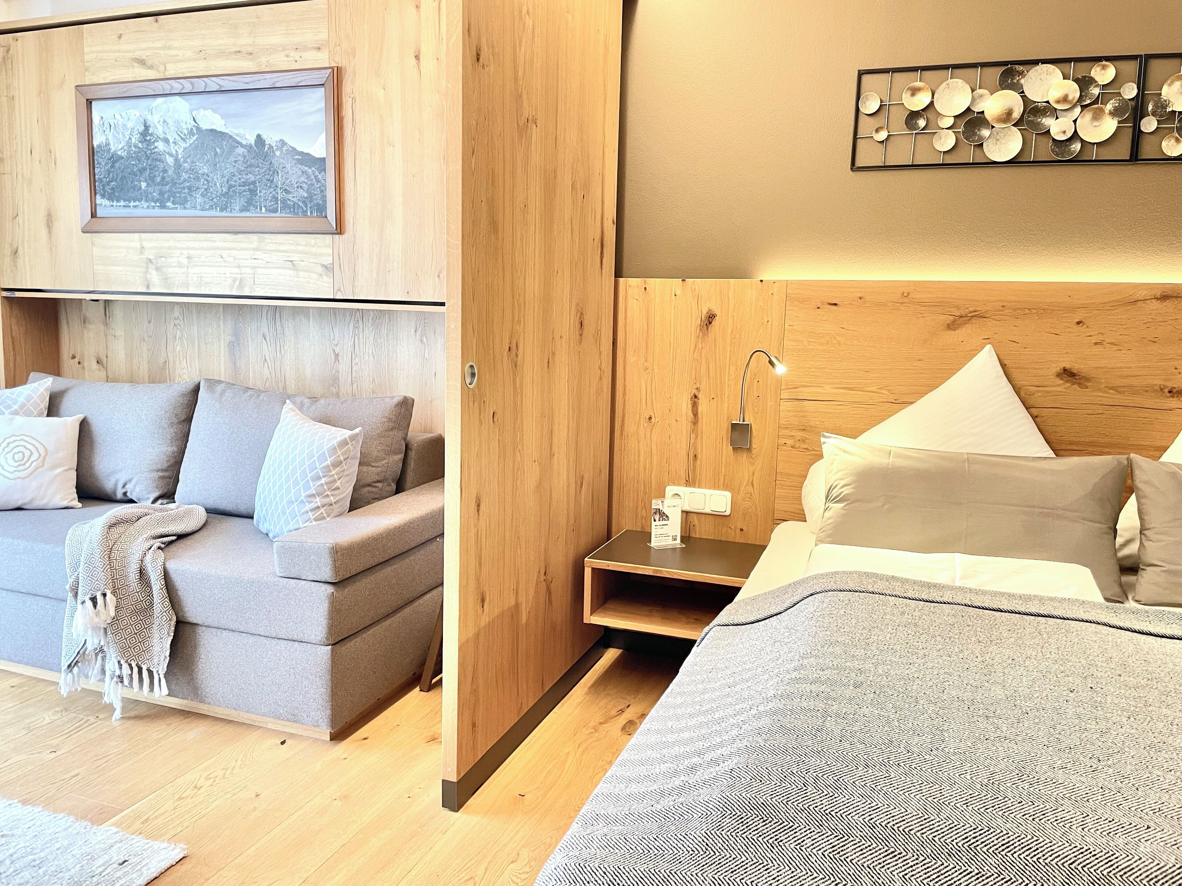 A modern hotel room with a wooden accent wall, a framed picture, a bedside table with a lamp, and a bed with a patterned bedspread.