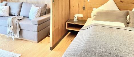 A modern hotel room with a wooden accent wall, a framed picture, a bedside table with a lamp, and a bed with a patterned bedspread.