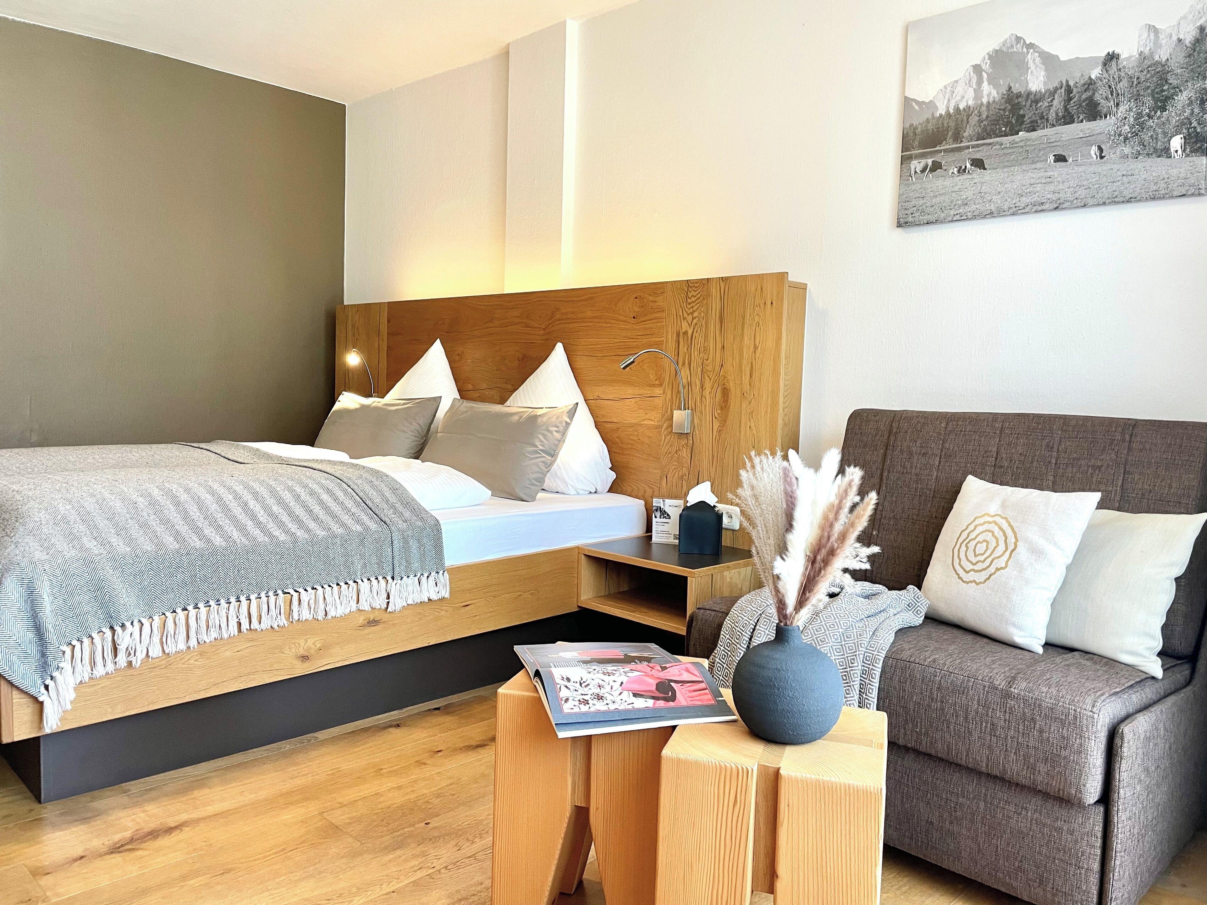 A modern hotel room with a wooden headboard bed, a grey patterned bedspread, a grey throw blanket, a wooden nightstand with a lamp, a magazine, a vase with dried flowers, and a grey sofa.