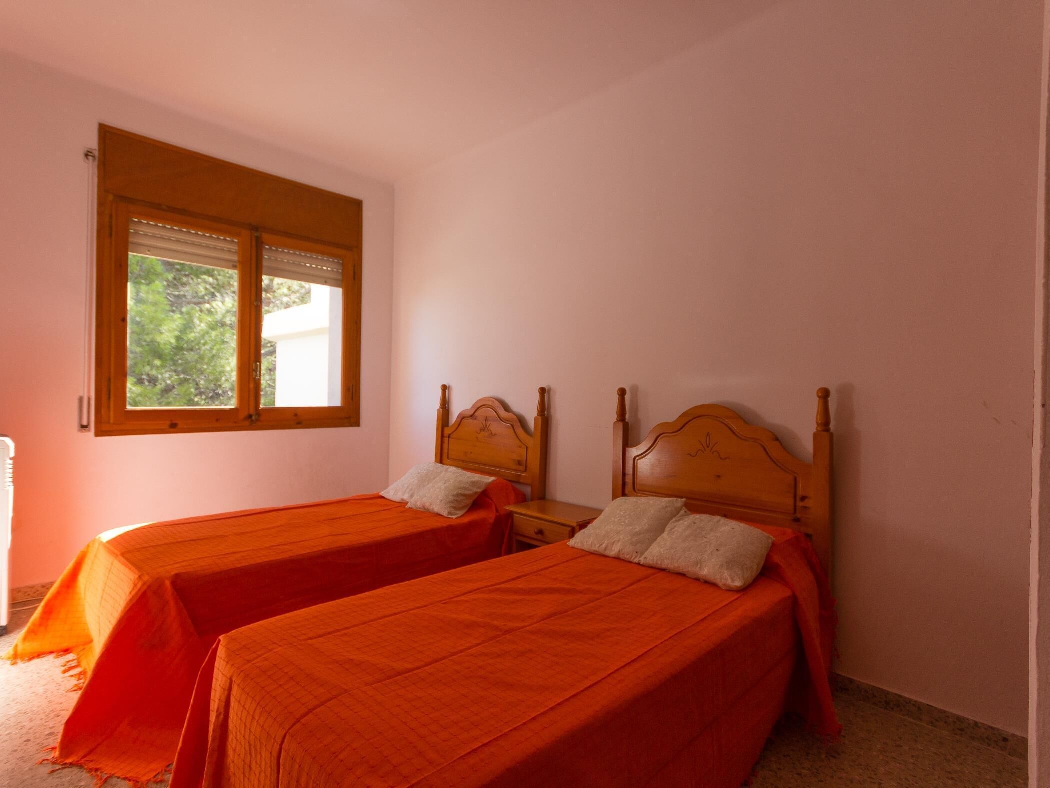 A room with two single beds, a window with blinds, and a view of greenery outside.