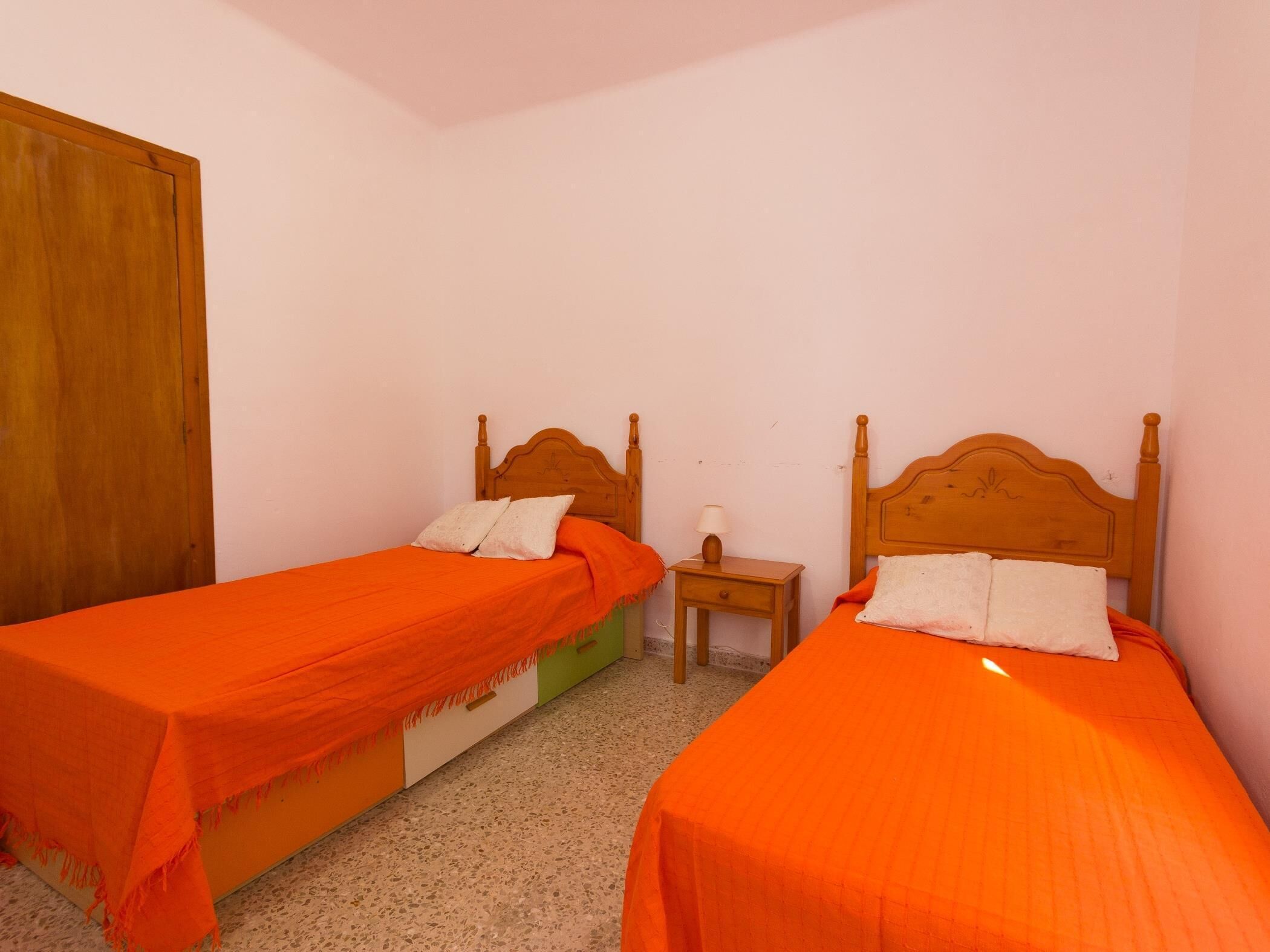 Une chambre avec deux lits simples, chacun doté d’une tête de lit en bois et d’une literie orange. On y trouve une petite table de chevet avec une lampe et un tiroir.