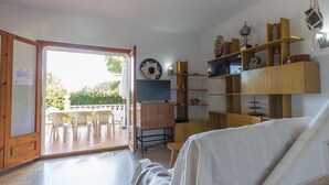 Ein Zimmer mit einem Holzkasten, einem Fernseher, Regalen mit Dekoration und einem Essbereich mit Stühlen, sowie einer Aussicht durch eine offene Tür.
