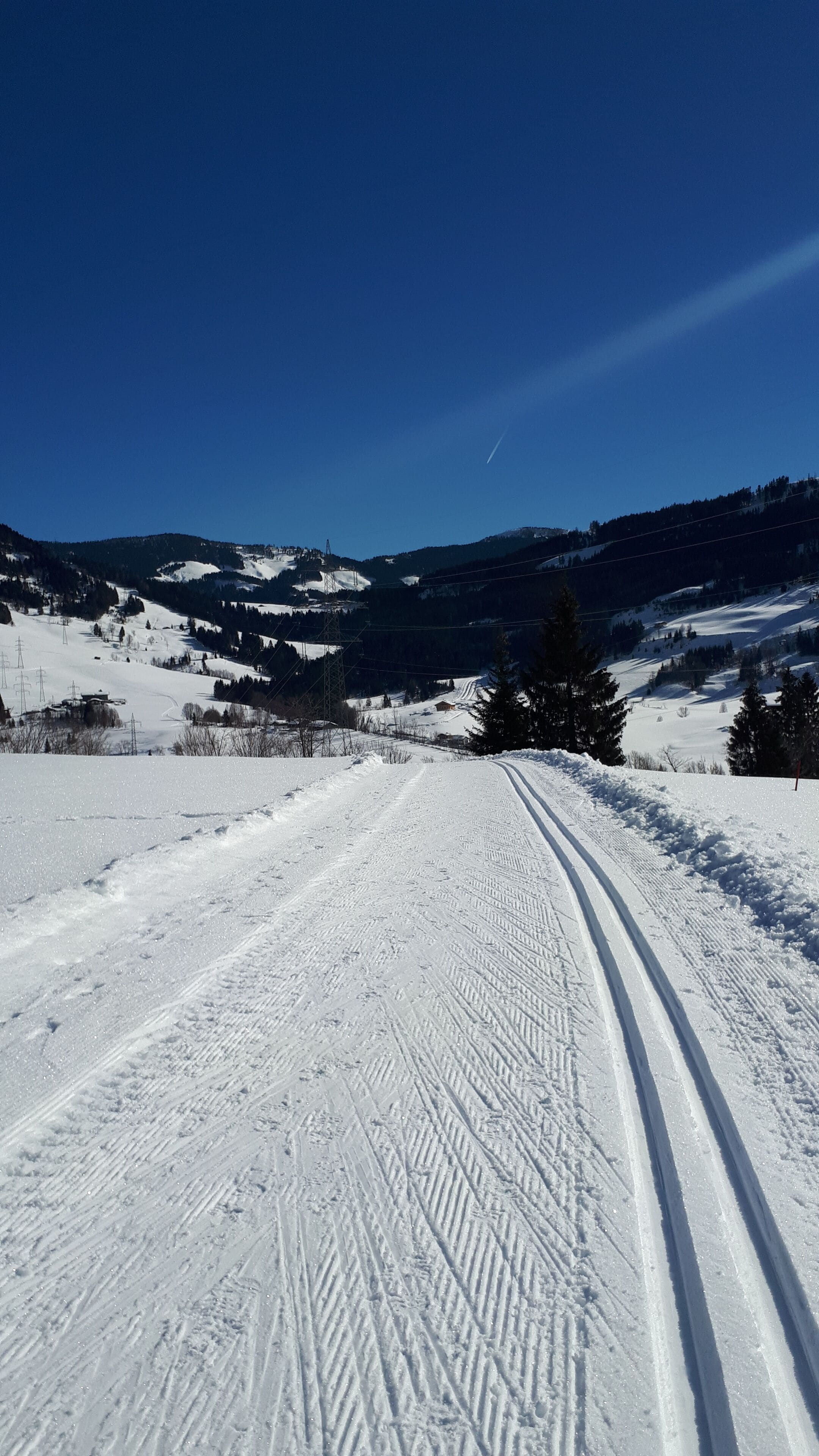 A snow-covered road with tire tracks, surrounded by forested hills and a clear blue sky.