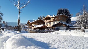A large wooden chalet with multiple balconies and a covered patio area, surrounded by snow-covered trees and a clear blue sky. - Hotel Sonnenlicht (Maria Alm am Steinernen Meer)