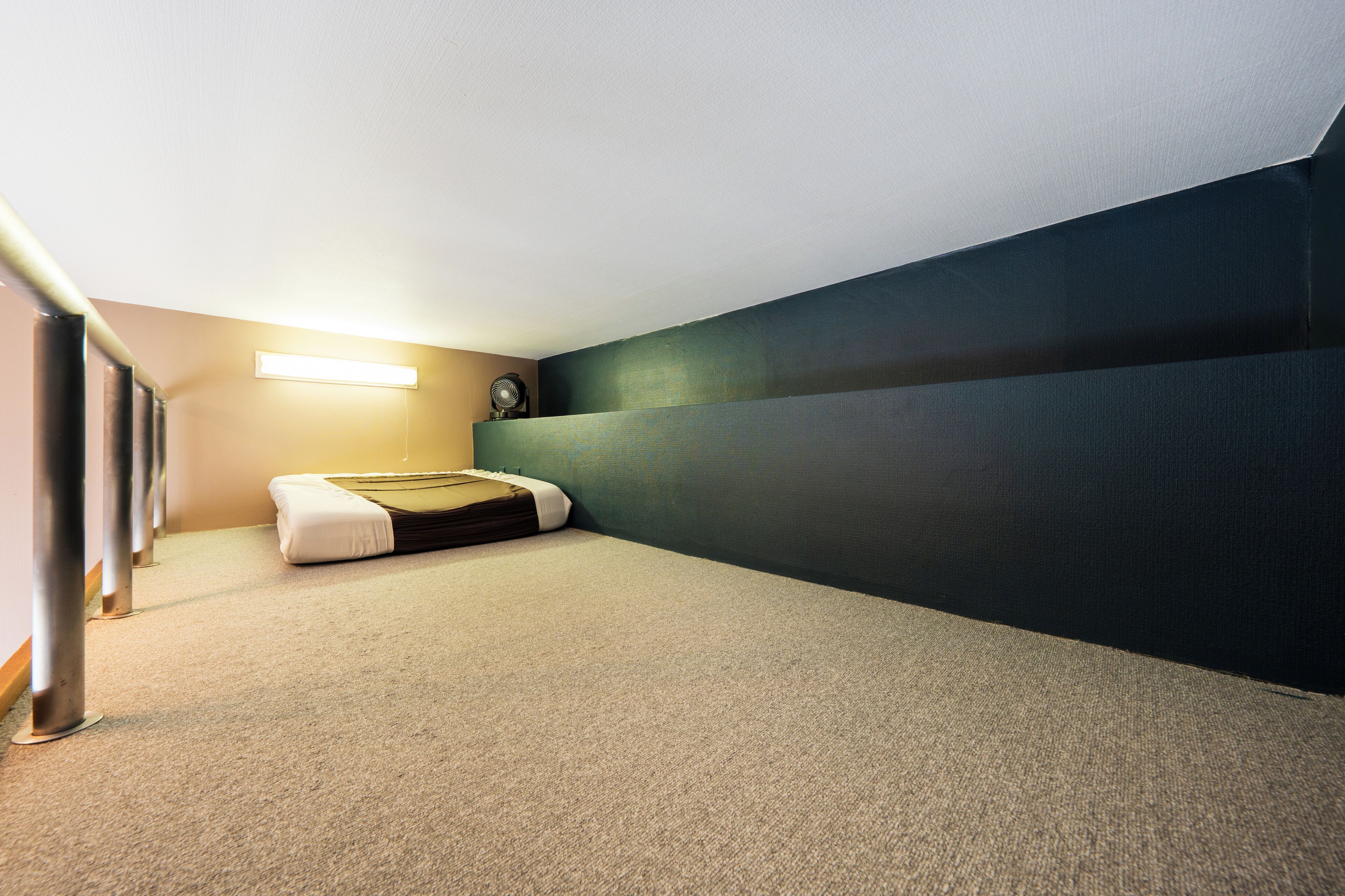 A small, carpeted room with a single bed, a wall-mounted clock, and a ceiling light.