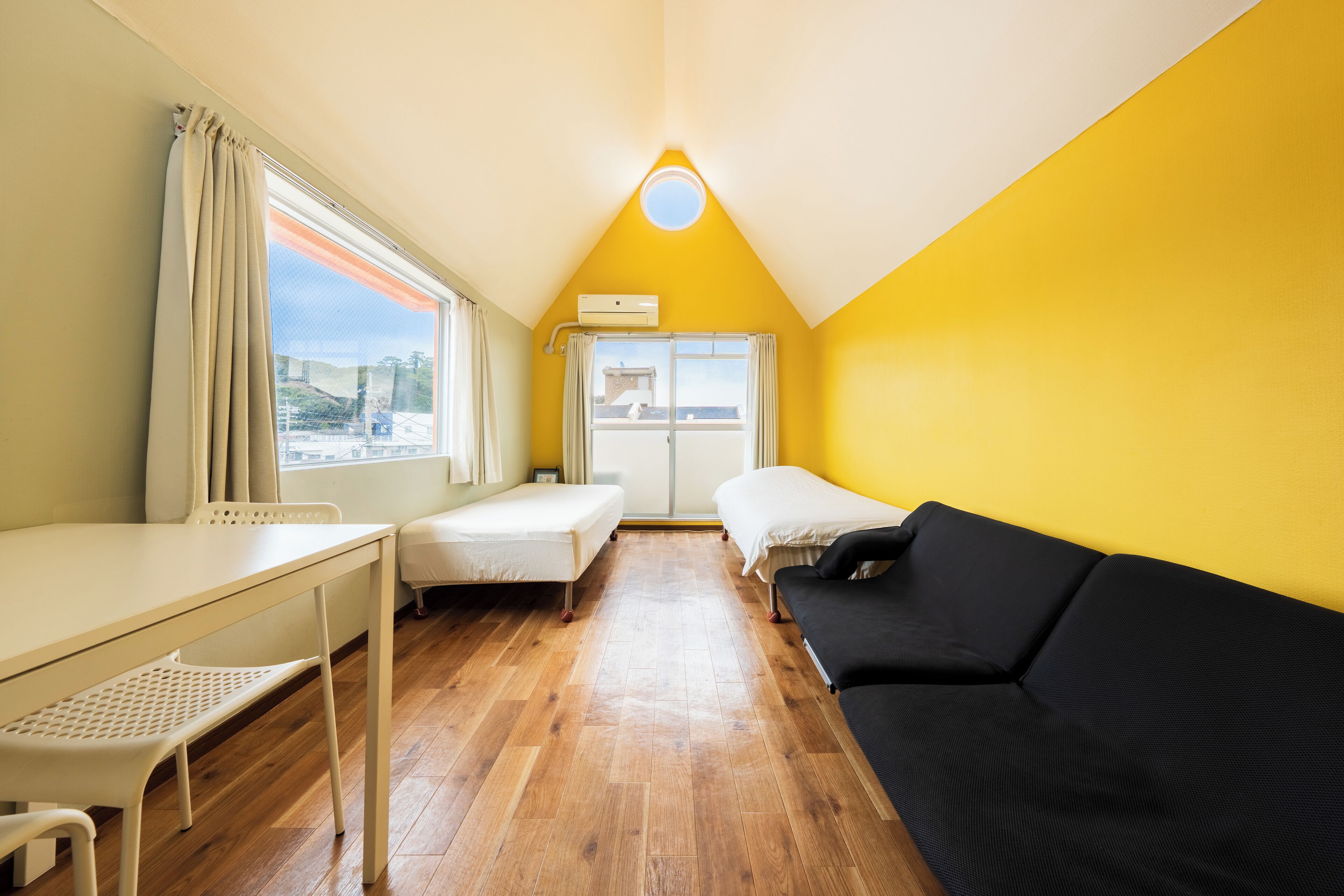 A small, bright room with a wooden floor, a white desk, a black sofa, and two beds.
