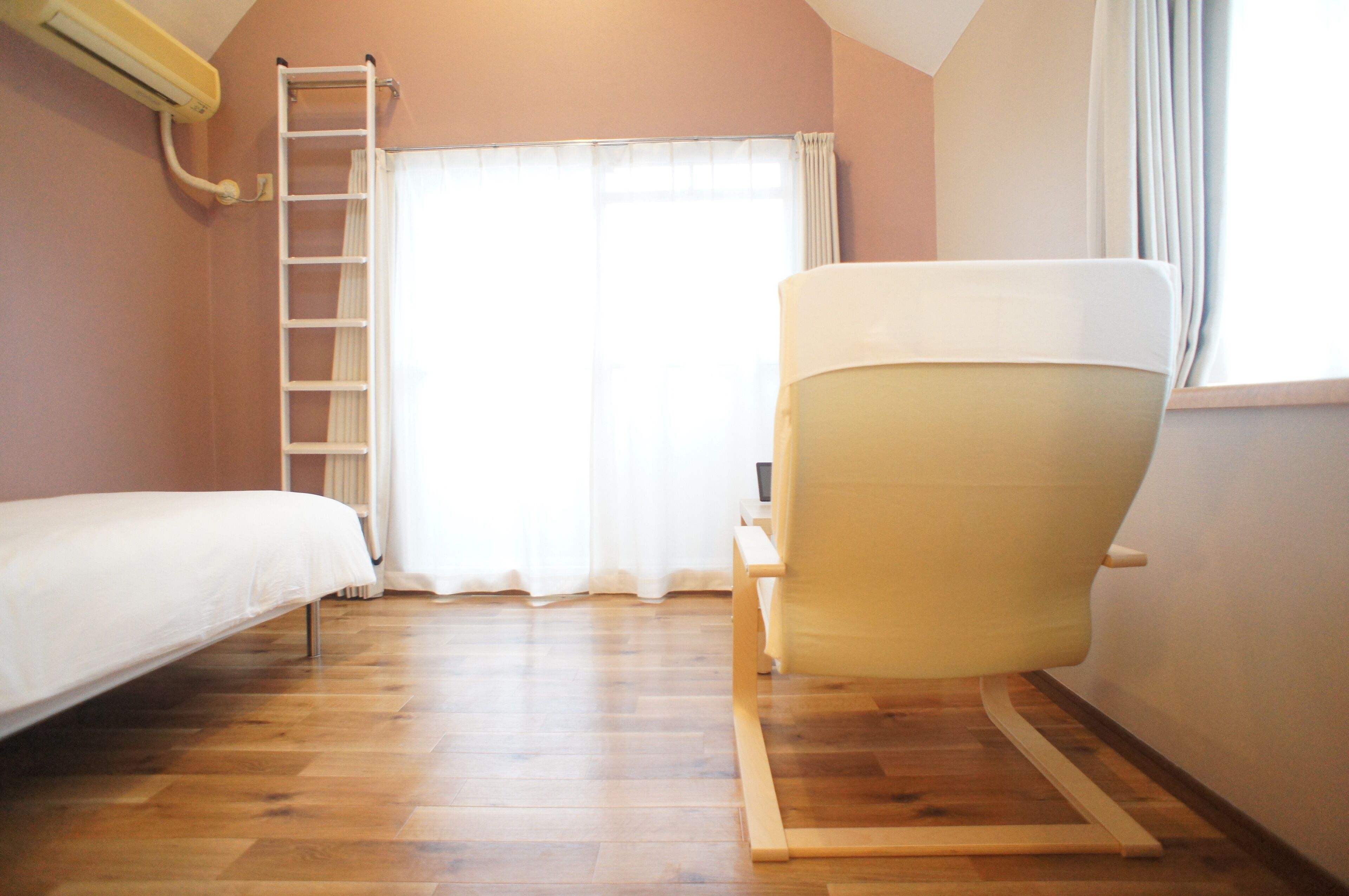A bedroom with a wooden floor, a bed, a chair, a ladder, and a window with curtains.