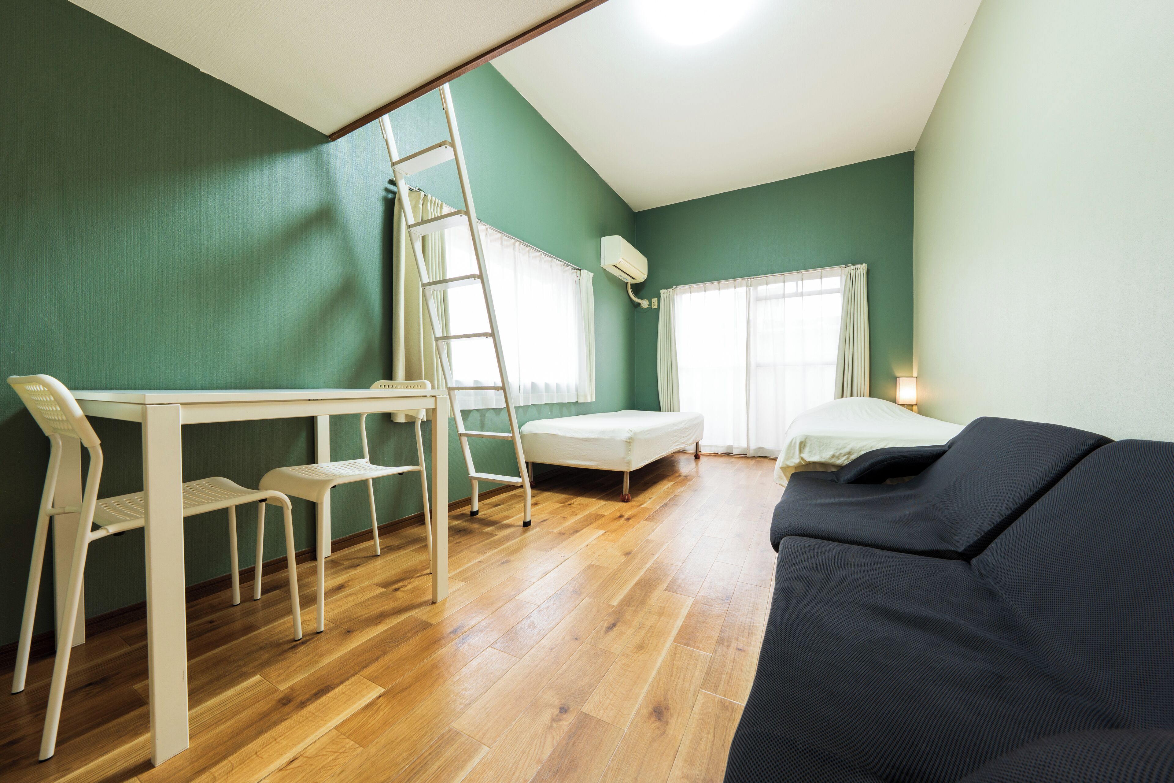 A room with a wooden floor, a white desk with two chairs, a black sofa, a ladder, a bed, and a window with curtains.