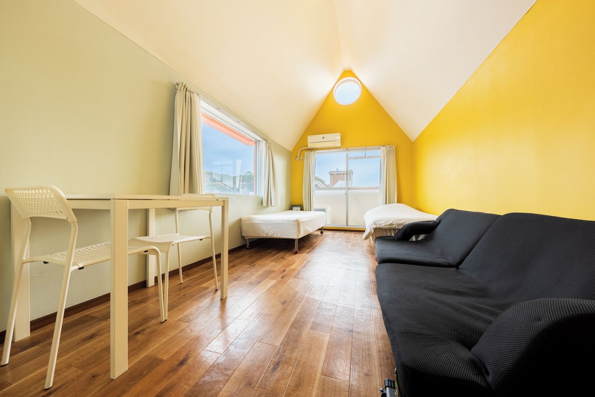 A small, bright room with a wooden floor, a black sofa, a small table, and a white chair.