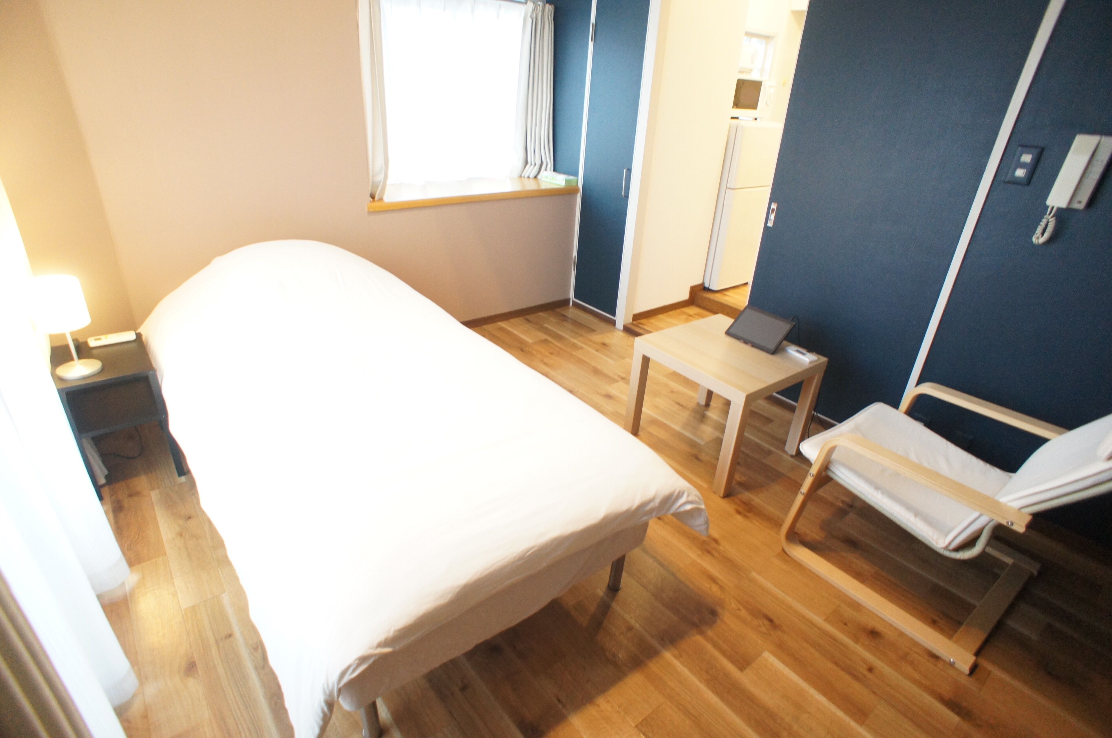 Une chambre d’hôtel avec un lit, une petite table, une chaise, une table de chevet, une fenêtre avec des rideaux et un mur bleu.