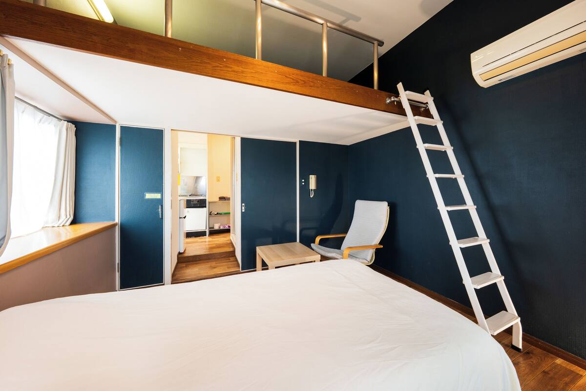 A bedroom with a large bed, a chair, a ladder, and a view of a kitchen area.